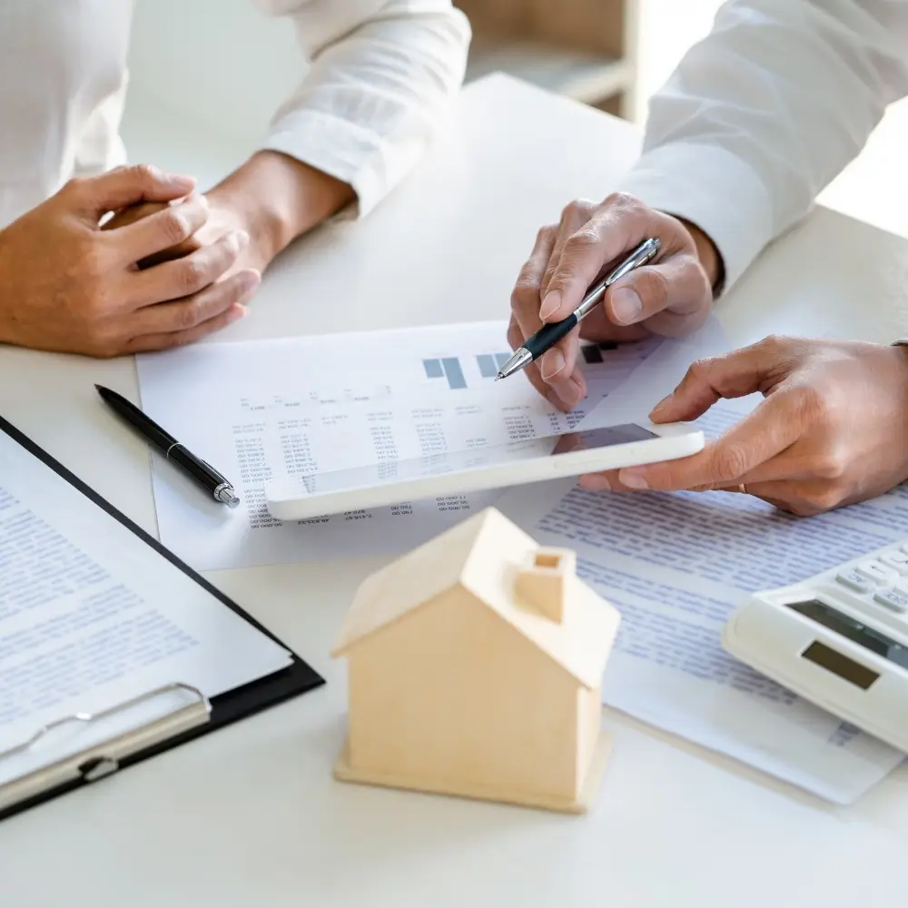 Two people review financial documents, a wooden house, and calculator—discussing purchase home loans at the desk. - Brad Hamblen Home Loans - Citywide Mortgage Two people review financial documents, a wooden house, and calculator—discussing purchase home loans at the desk.