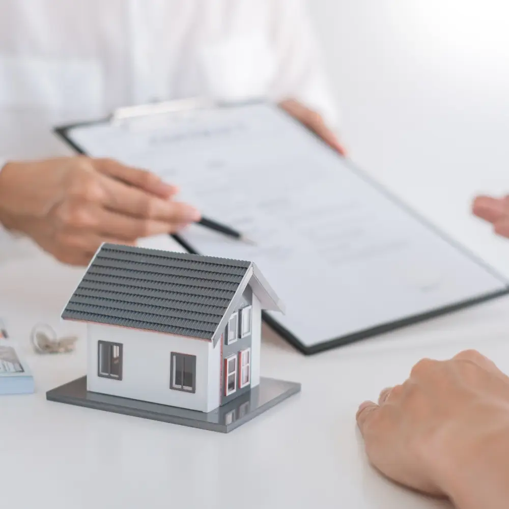 A person points to paperwork by a model house, indicating a real estate transaction with a mortgage lender.