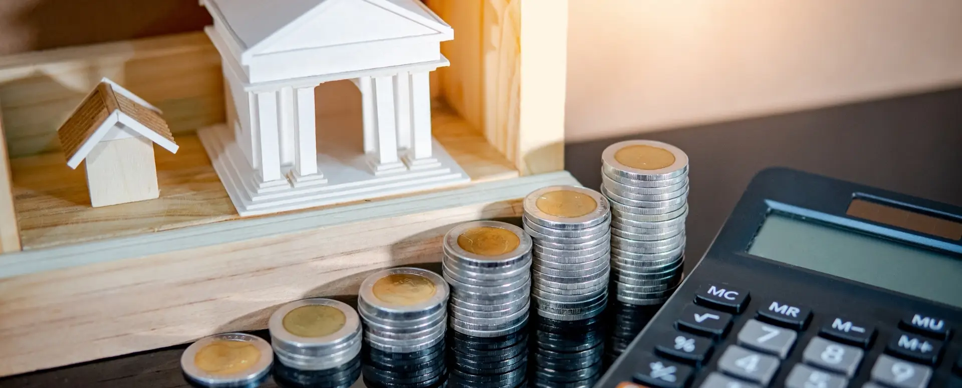 Stacks of coins, a calculator, and a bank model symbolize mortgage lender options and home loans on a reflective surface.