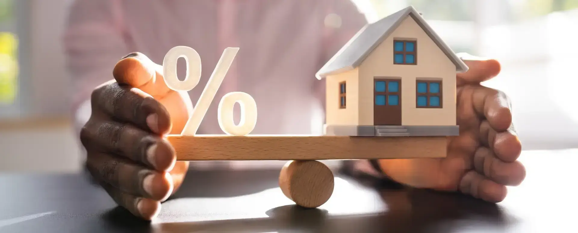 Hands balance a wooden plank with a house model and percent sign, symbolizing mortgage refinance or interest rates.