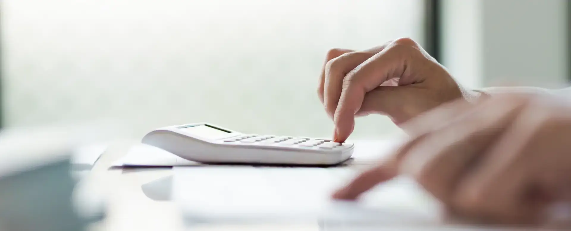 A hand presses buttons on a white calculator, perhaps calculating a mortgage refinance at a desk with blurred papers.