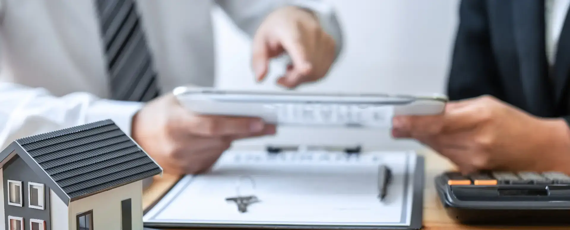 Two people review documents on a tablet, discussing home loans with a model house and clipboard in the foreground.