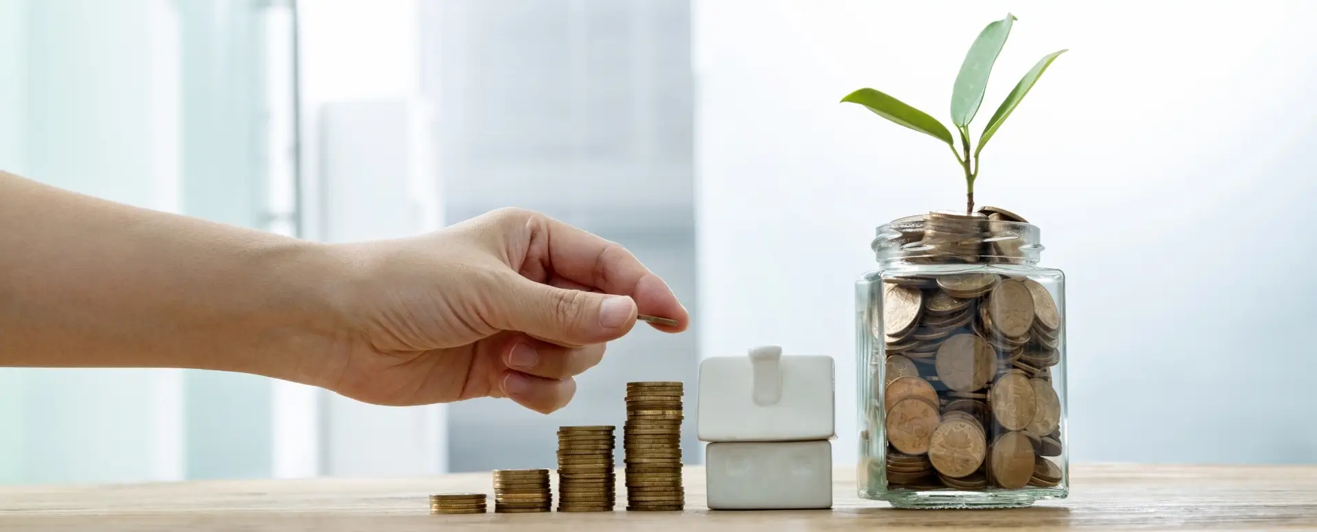 A hand stacking coins beside a toy house and jar of coins with a plant—perfect for illustrating home loans.
