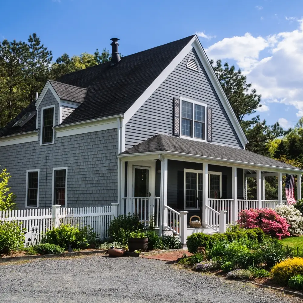 A gray house with a porch and garden, perfect for home loans, sits by trees under a blue sky with clouds.