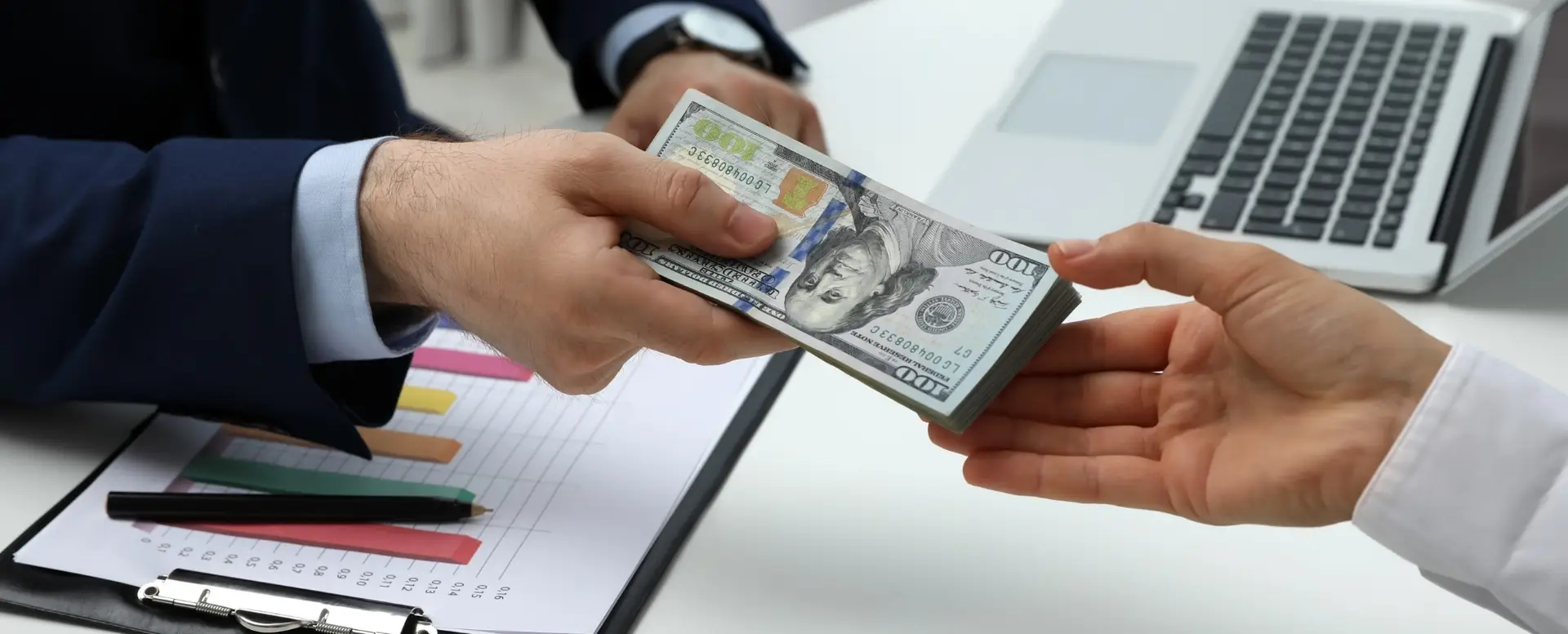 A mortgage lender hands a stack of US dollars to a first-time home buyer near a laptop and clipboard with charts.