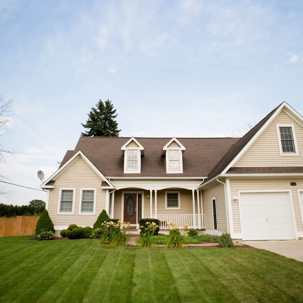 A beige suburban house perfect for first-time home buyers, with a porch, garage, and neat lawn under a blue sky.