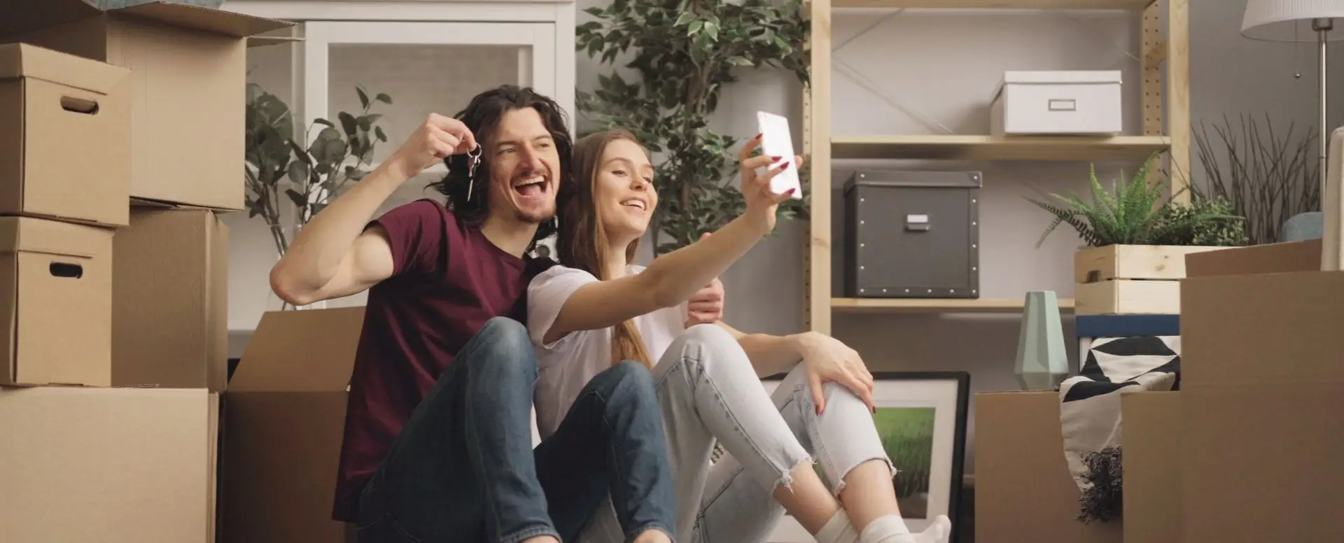 Smiling couple with house keys and moving boxes snaps a selfie, celebrating their new home and mortgage lender.