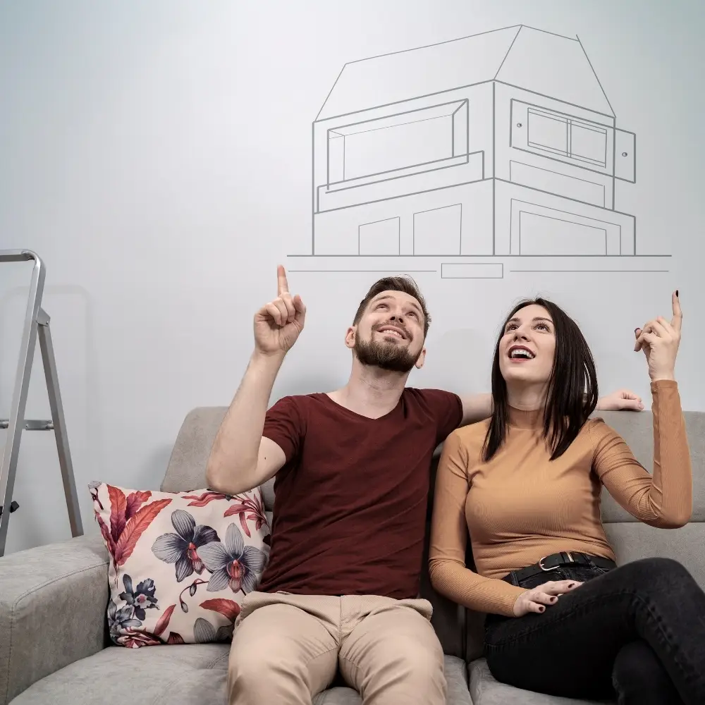 A smiling couple sits on a couch, pointing at a house blueprint—excited first-time home buyers discussing home loans.