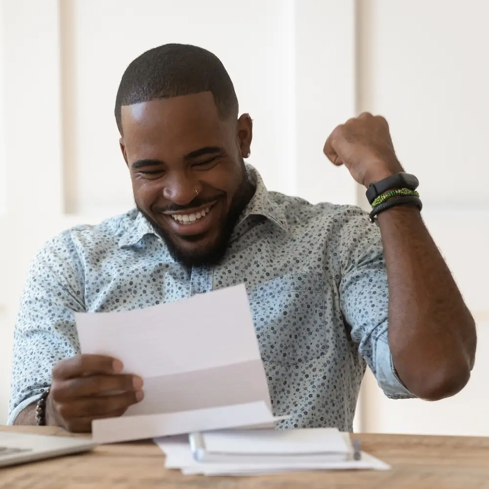 Smiling man sitting at a table, holding a letter from his mortgage lender and raising his fist in excitement.