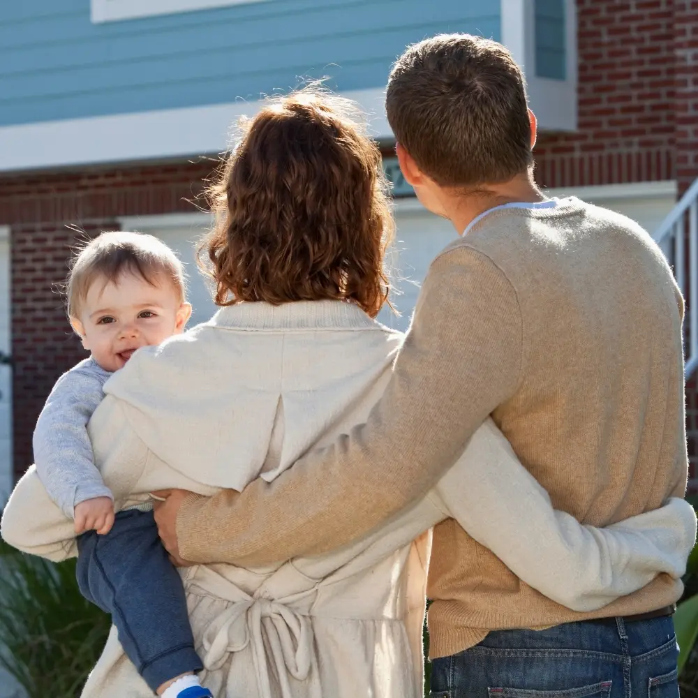 A couple stands with their backs to the camera, holding a smiling baby—first-time home buyers outside a new house.