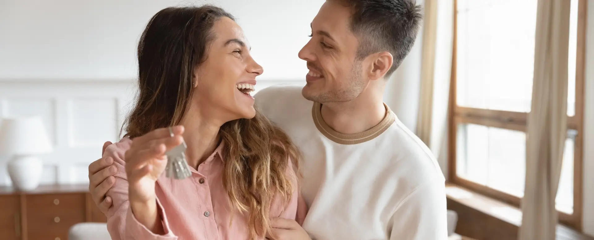 A smiling couple embraces indoors with keys, celebrating their new home as first-time home buyers.