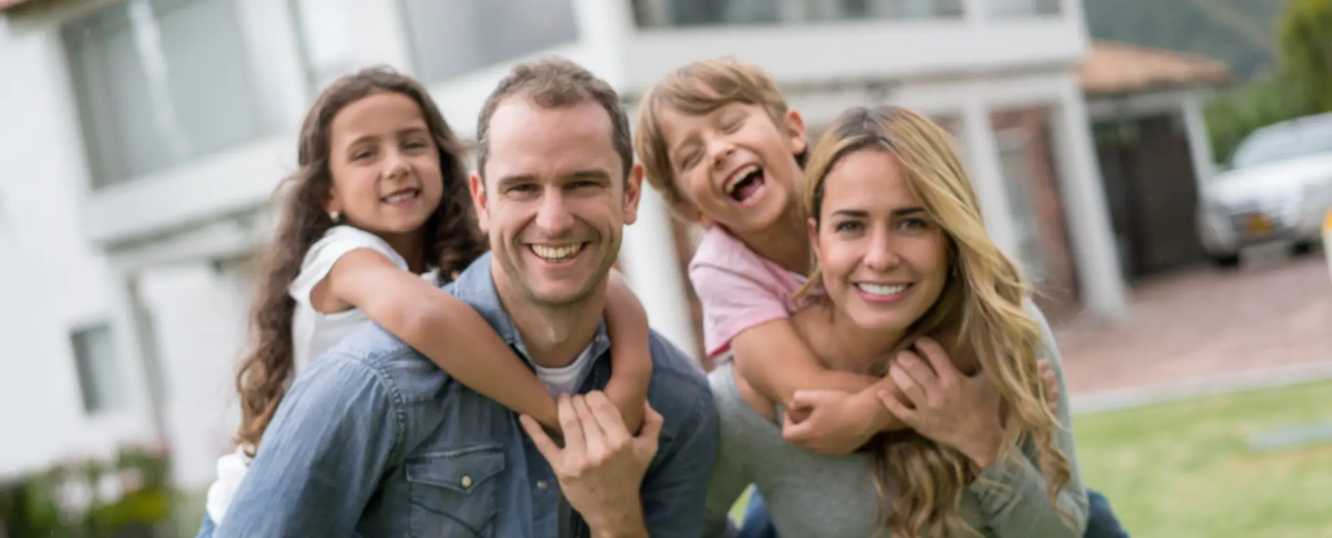 Smiling couple gives piggyback rides to laughing kids outside their new home—perfect for first-time home buyers.