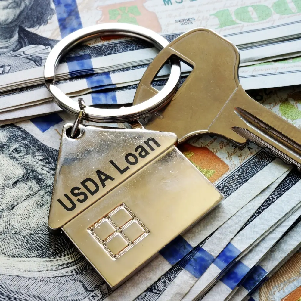 A house-shaped USDA Loan keychain for first-time home buyers rests with keys on a stack of hundred dollar bills.