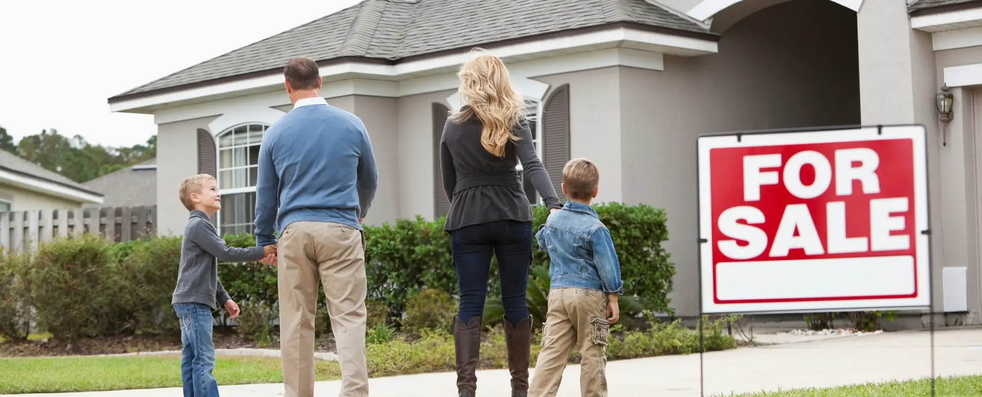 A family of four stands outside a gray house with a For Sale sign, ready for purchase home loans as first-time home buyers.