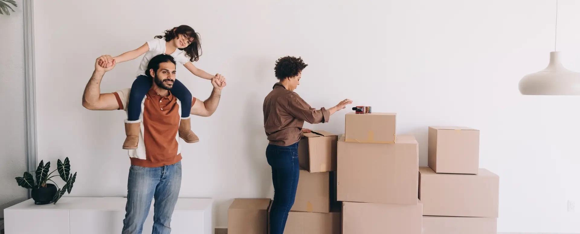 A man carries a smiling girl as a woman stacks boxes—first-time home buyers enjoying their new space.