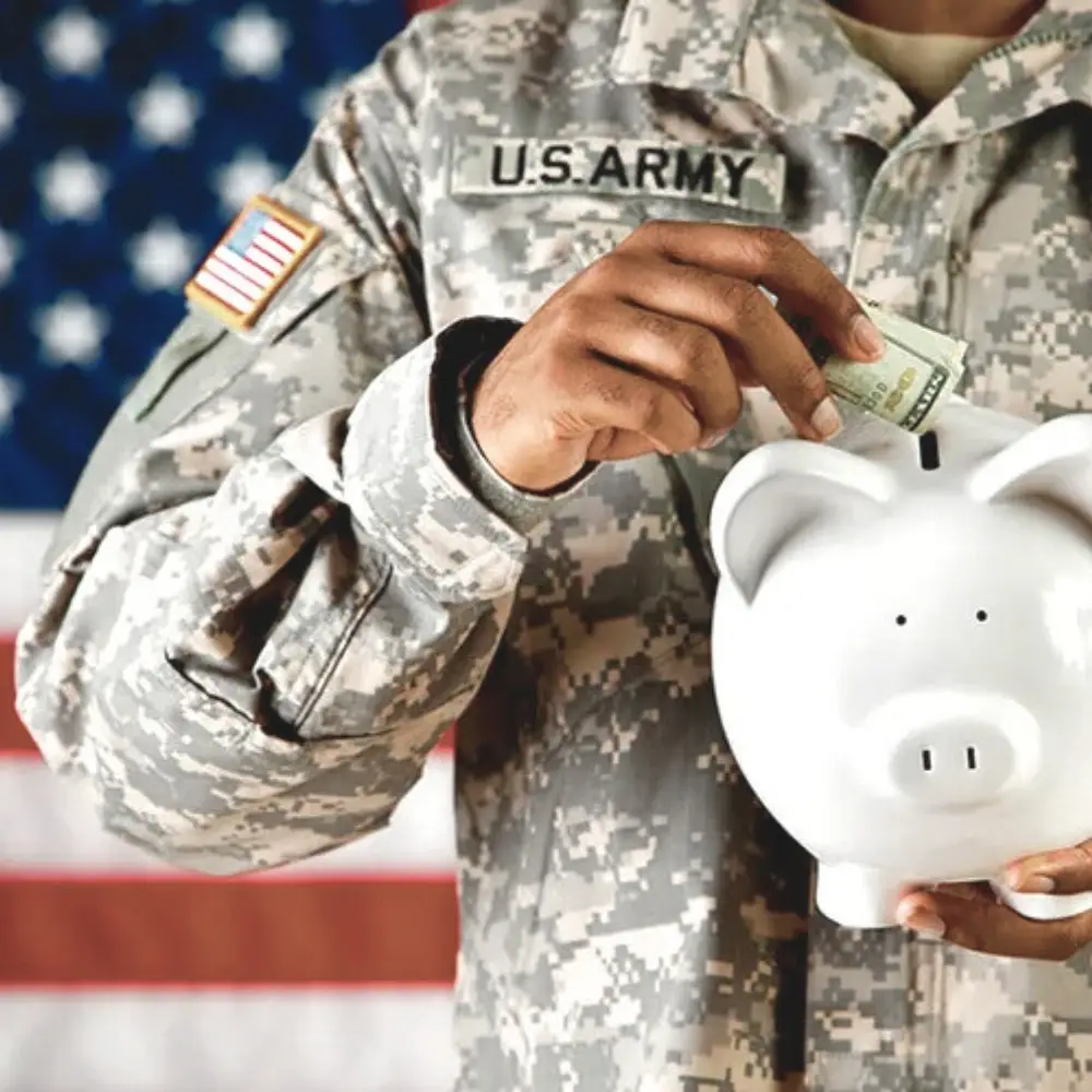 A U.S. Army soldier puts a dollar bill into a white piggy bank, symbolizing saving for home loans, with an American flag behind.