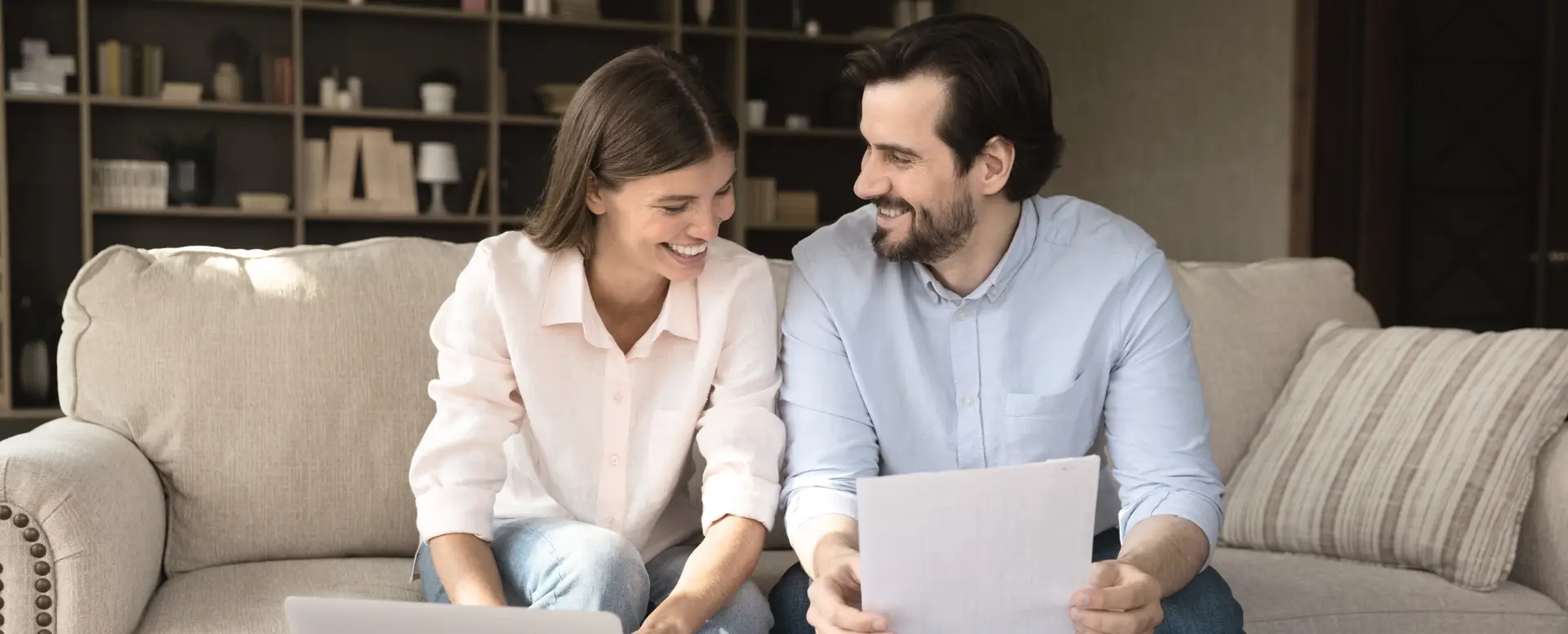 Smiling couple sitting on a couch, reviewing construction loan papers together, with a laptop in front of them.