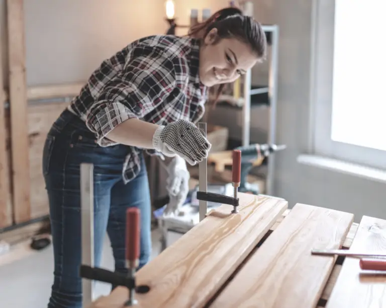 Smiling woman in plaid shirt clamps boards, learning carpentry—perfect inspiration for first-time home buyers.