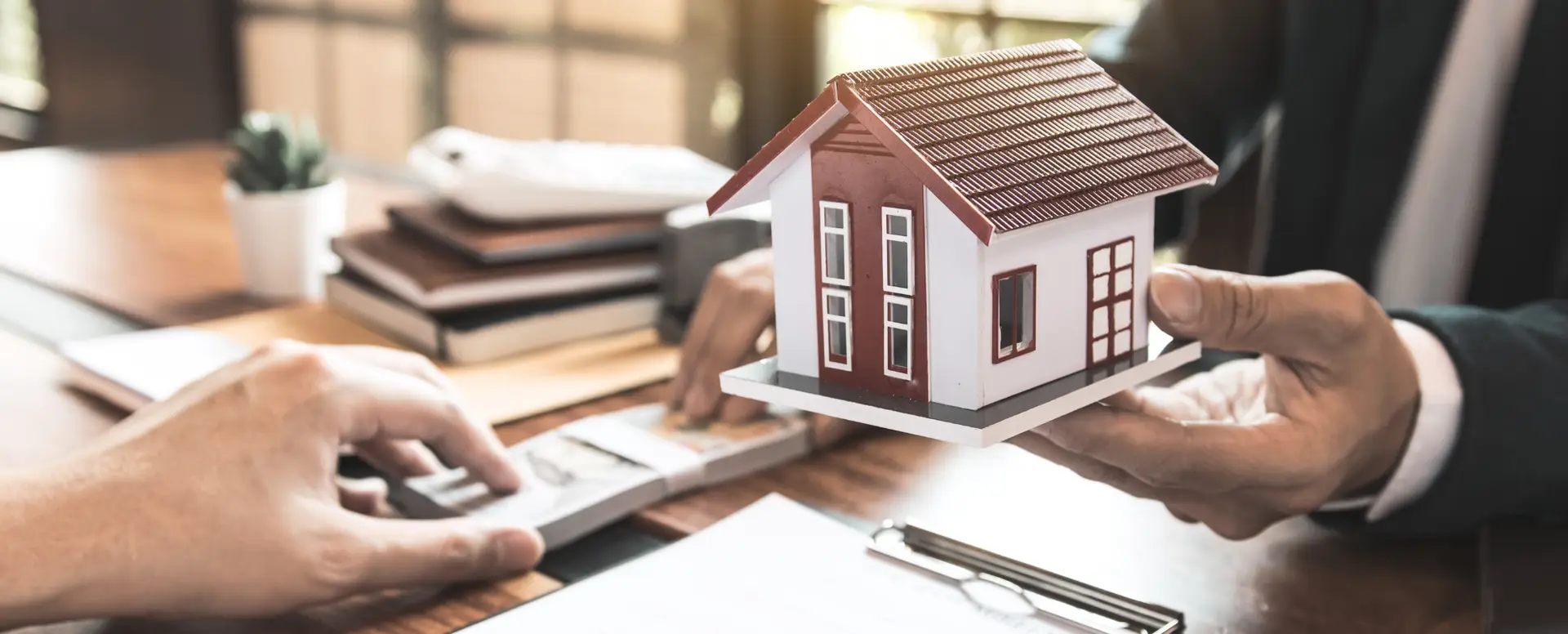 A person hands a model house to another across a desk, discussing purchase home loans with documents and a calculator nearby.