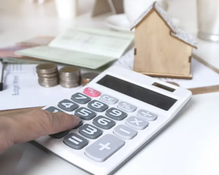 A hand presses calculator buttons near coins, papers, and a house model—perfect for managing purchase home loans.