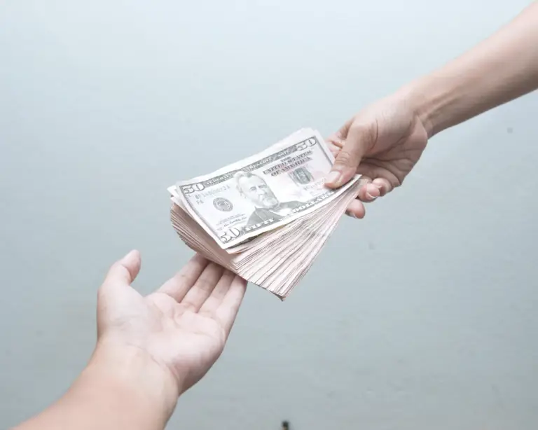 One hand gives a stack of fifty dollar bills to another, symbolizing a Mortgage Refinance, against a plain background.