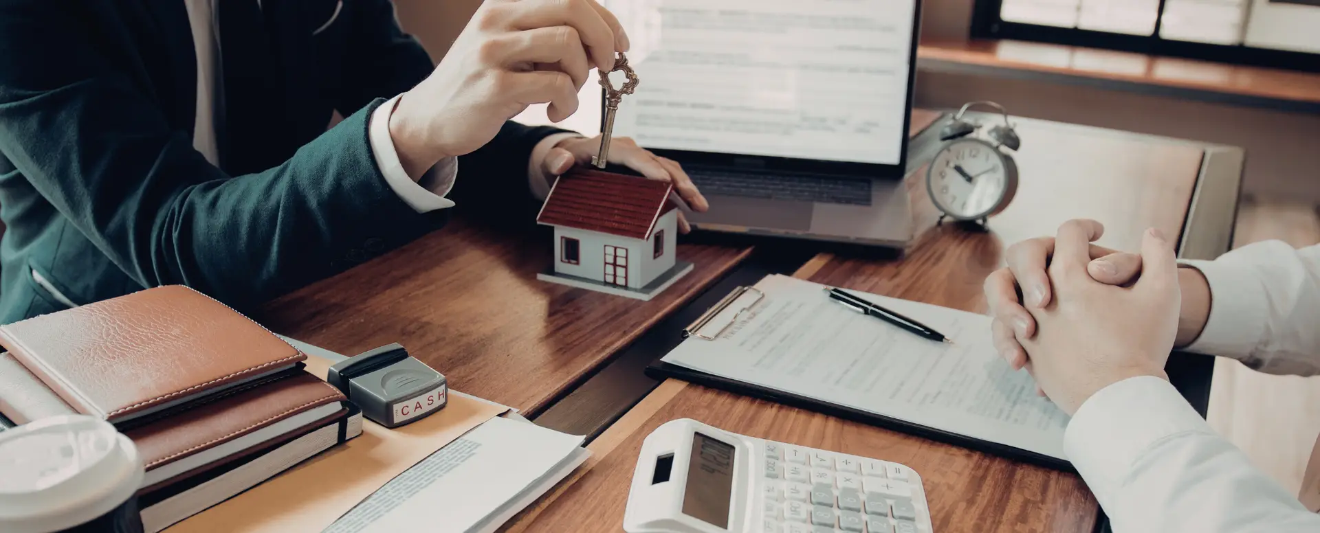 A mortgage lender hands a house key to first-time home buyers over documents and a model house on a wooden desk.