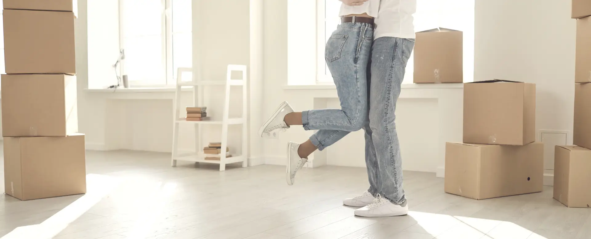 A couple in jeans embraces among moving boxes, celebrating with joy after securing a purchase home loan.