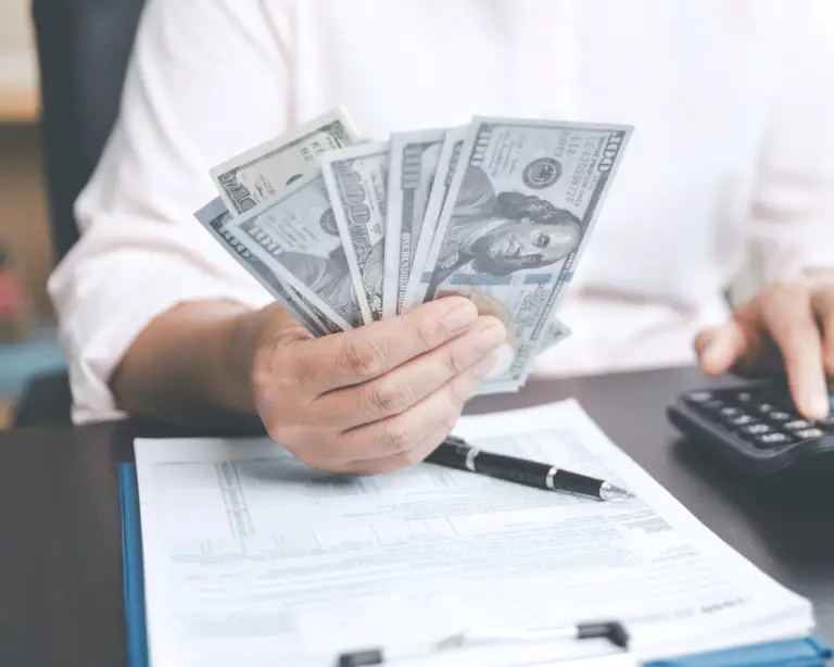 Person holding US dollar bills, with mortgage lender documents, clipboard, and calculator on the desk.