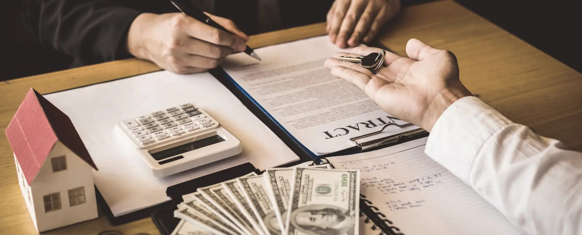 Two people signing a contract for home loans with house model, money, calculator, and keys on the table.
