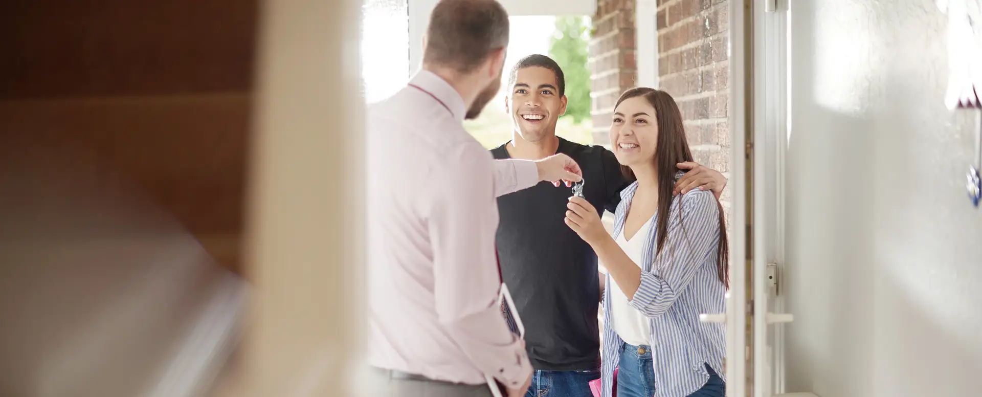 A mortgage lender hands keys to smiling first-time home buyers at the entrance of their new house.
