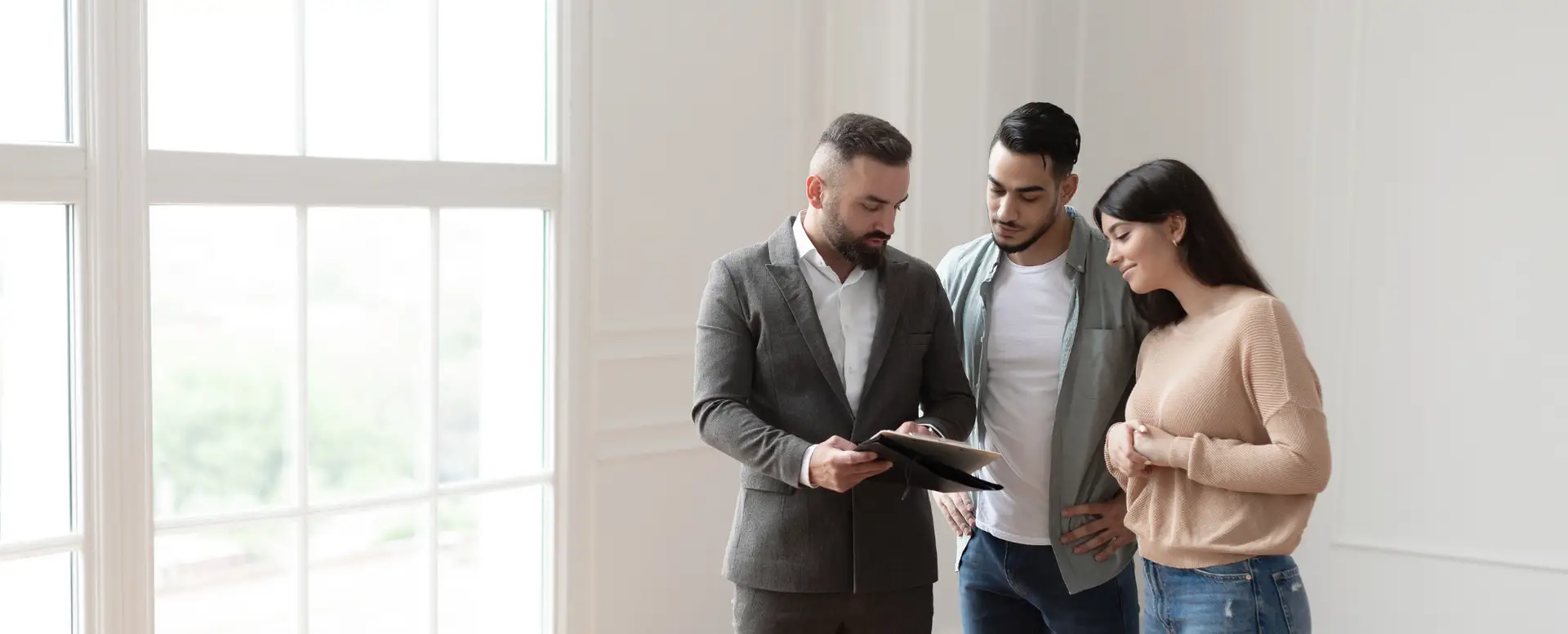 A real estate agent discusses home loans with a couple inside a bright, modern room with large windows.