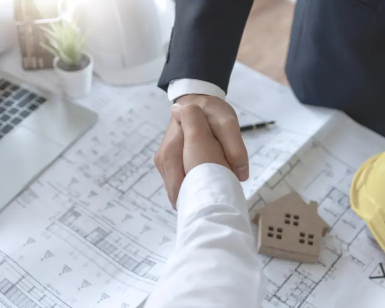 Two people in business attire shake hands over house plans—perfect for discussing construction loans or home loans.