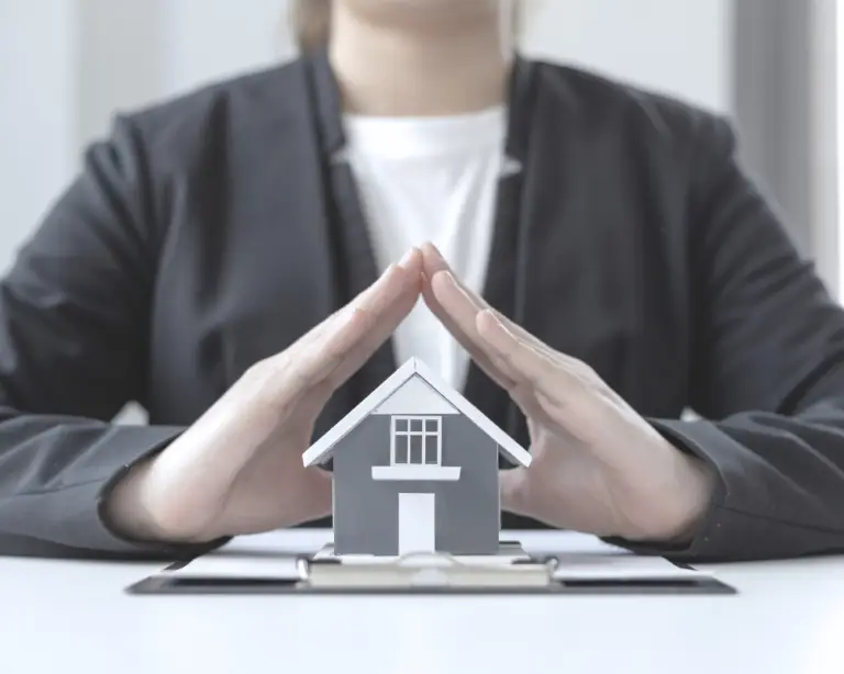 Person shielding a model house with hands, symbolizing protection for first-time home buyers or property insurance.