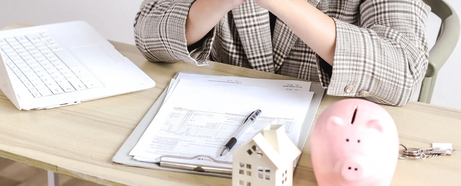 A person sits at a desk with documents, a pen, a laptop, and a small house model, planning for home loans.