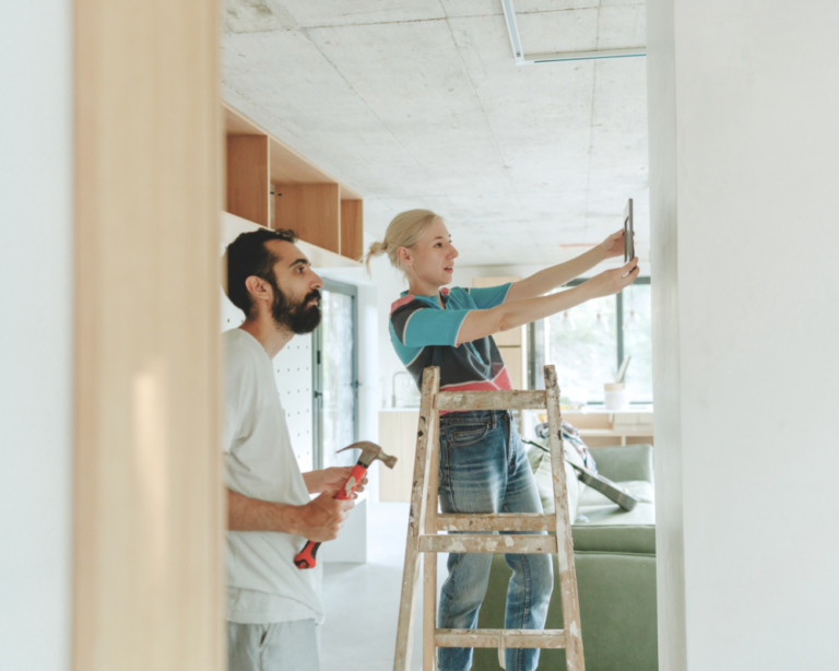 A woman on a ladder hangs a picture as a man holds tools—perfect teamwork for first-time home buyers.
