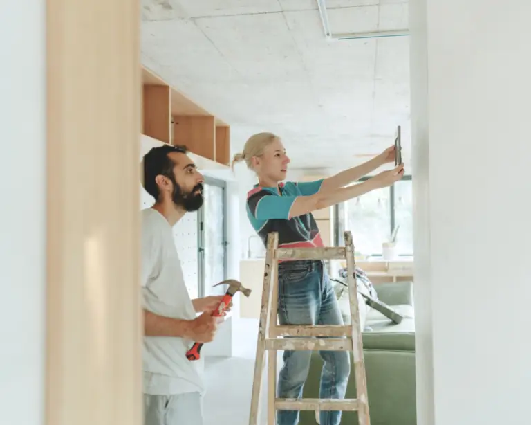 A woman on a ladder hangs a picture as a man holds tools—perfect teamwork for first-time home buyers.