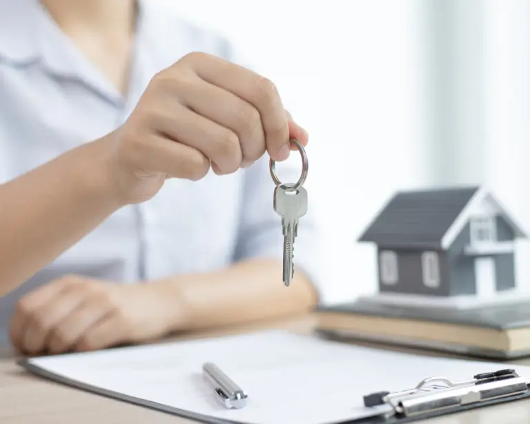 Person holding house keys over a desk with documents, a pen, and a mortgage lender's model house in the background.