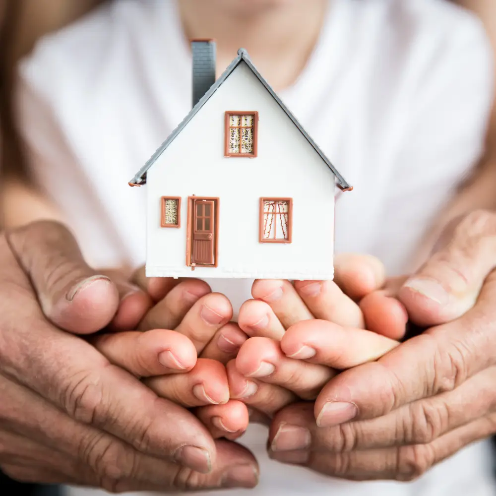 Several hands of different ages hold a model house, symbolizing family, home security, and the support of home loans.