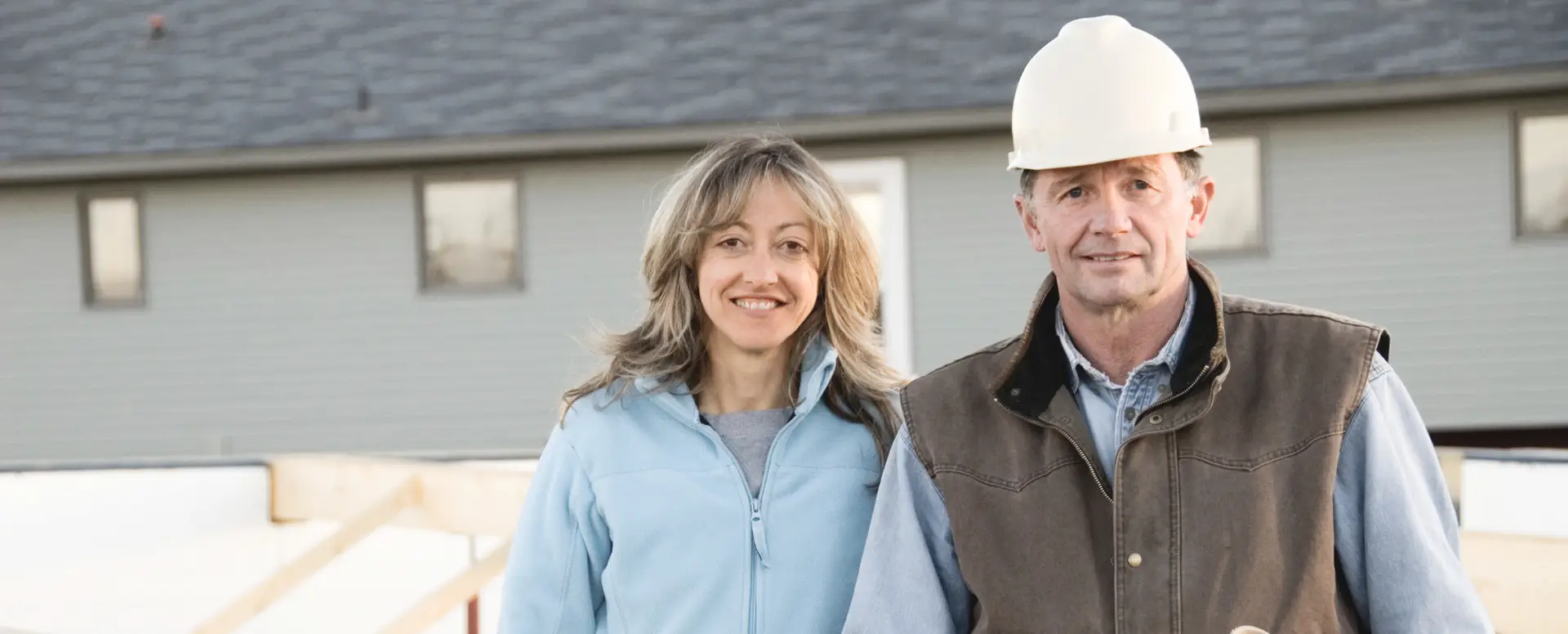 A woman and man in a hard hat smile outside a house under construction, discussing home loans.