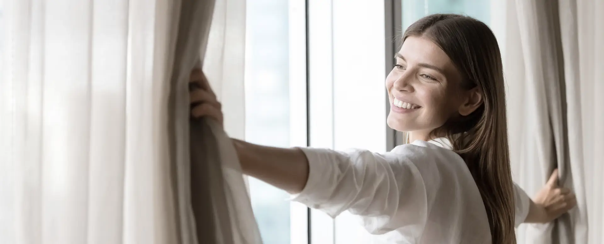 Smiling woman opens curtains to let sunlight into a bright room, perfect for first-time home buyers.