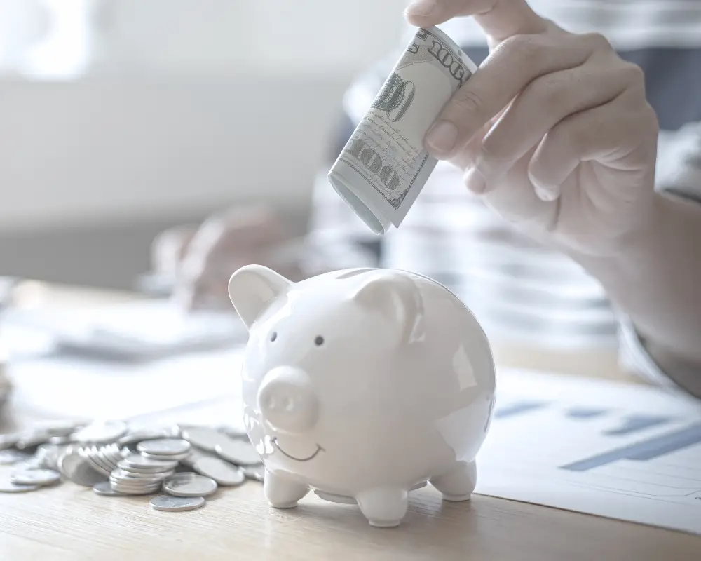 A person puts a rolled-up dollar bill into a piggy bank, saving coins for mortgage refinance or home loans.