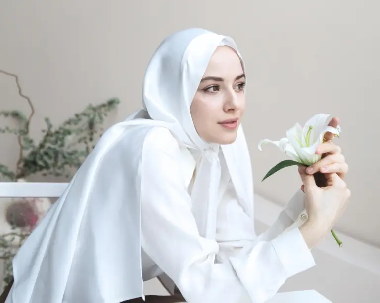 A woman in a white hijab smiles softly, holding a white lily—dreaming of home loans at a table with greenery behind her.