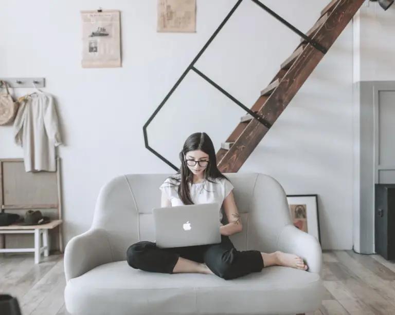 Woman sits cross-legged on sofa, researching mortgage refinance options on her laptop in a modern, minimalist living room.