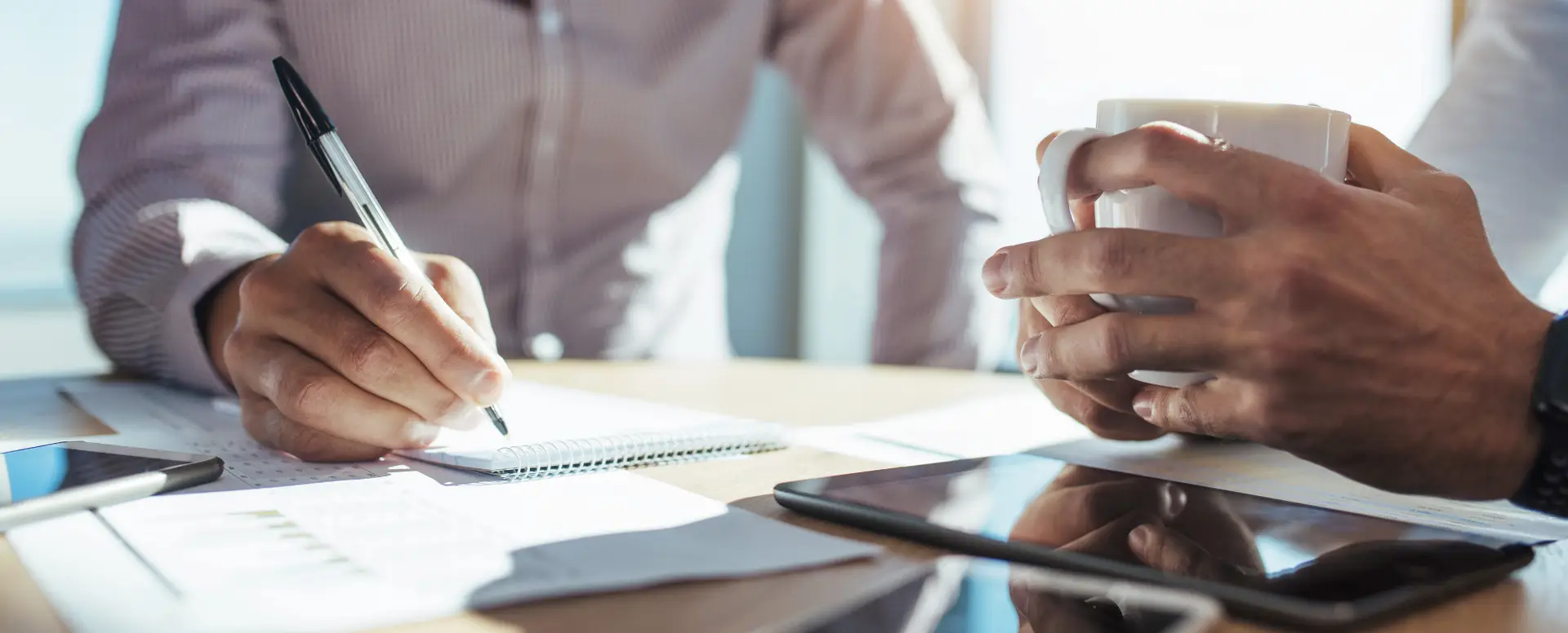Two people at a desk discuss home loans; one takes notes while the other holds a mug, tablet and papers nearby.
