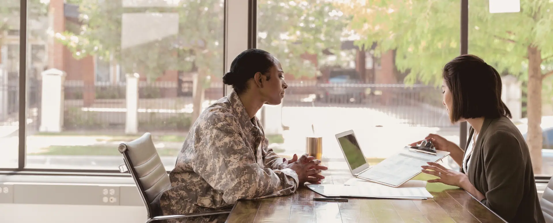 Two people, one in military uniform, discuss mortgage refinance documents at a bright office table with a laptop.