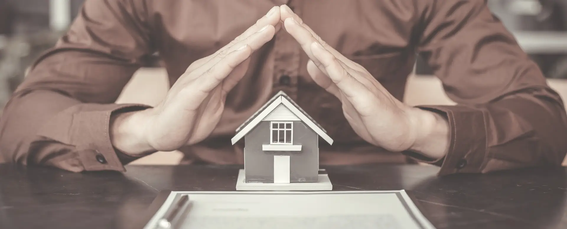 Person in a brown shirt sheltering a model house, highlighting home loans with a pen and document on the table.
