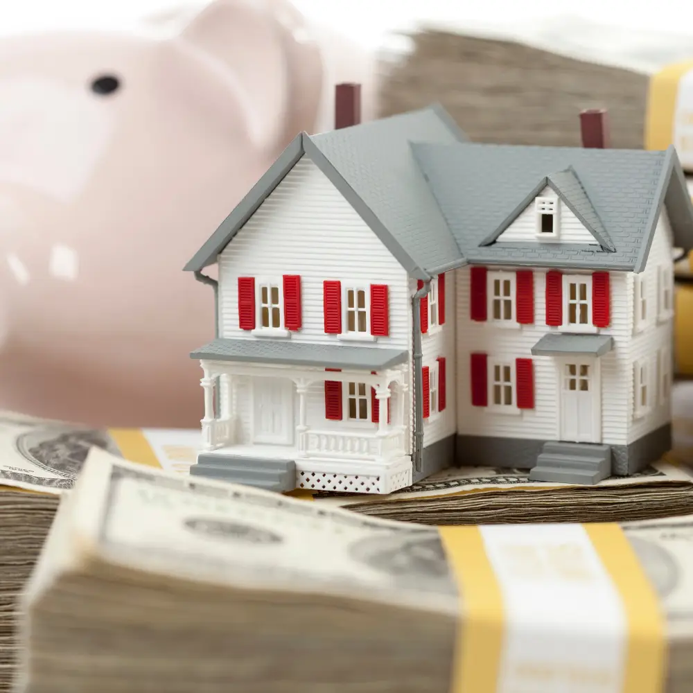 A model house sits on stacks of cash, symbolizing home loans for first-time home buyers, with a piggy bank behind.
