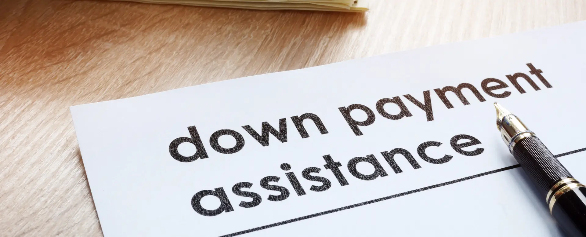 A paper labeled down payment assistance beside a pen, highlighting home loans on a wooden desk with documents in the background.