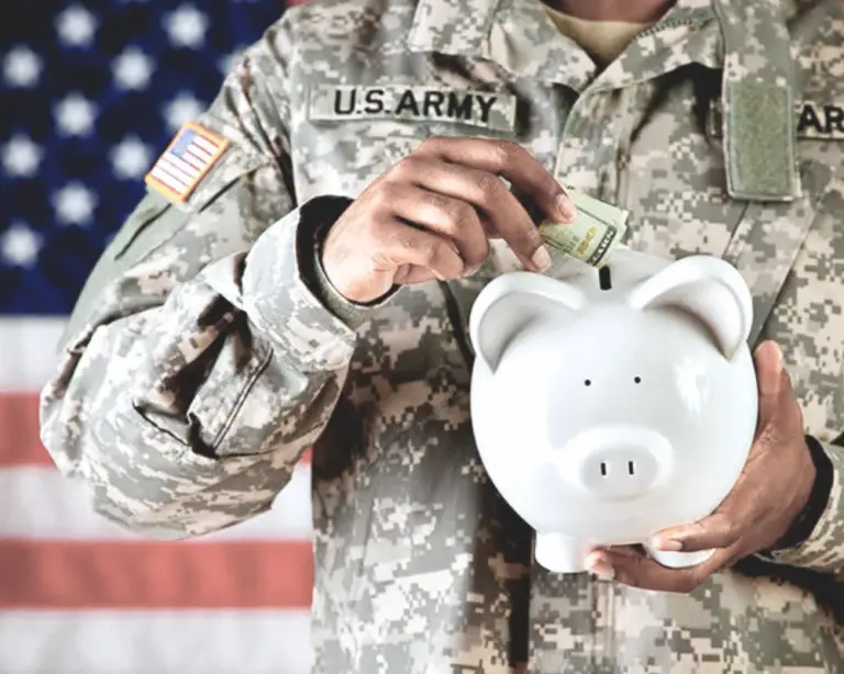 A person in U.S. Army uniform puts cash into a piggy bank, saving for home loans, with an American flag in the background.