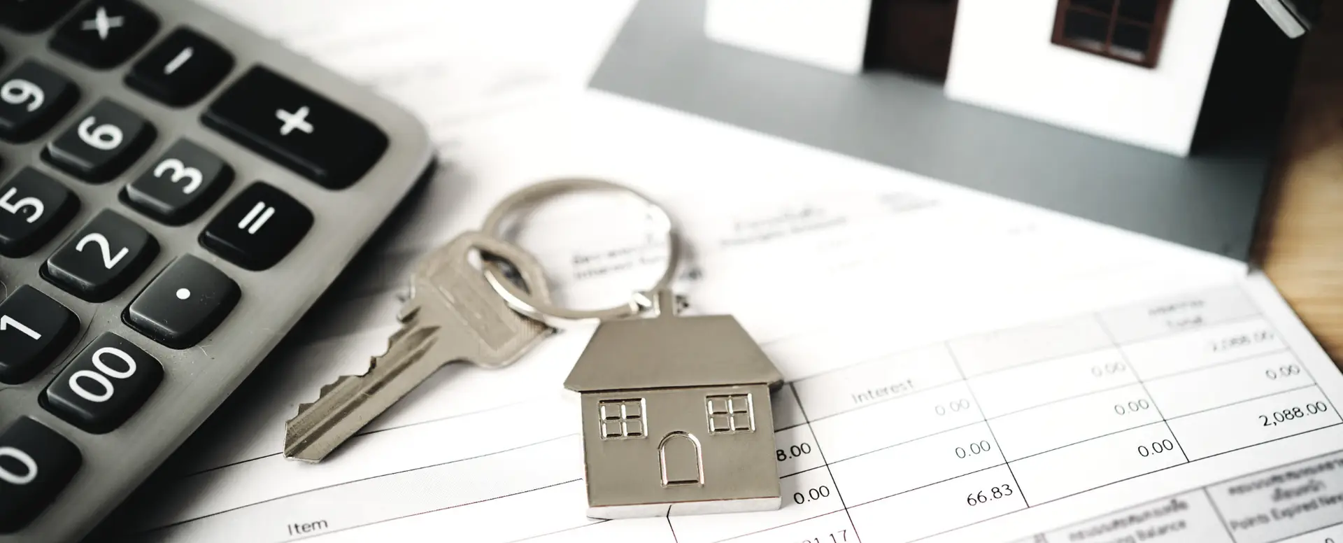 A house key with a house-shaped keychain on financial documents beside a calculator and mortgage lender model house.